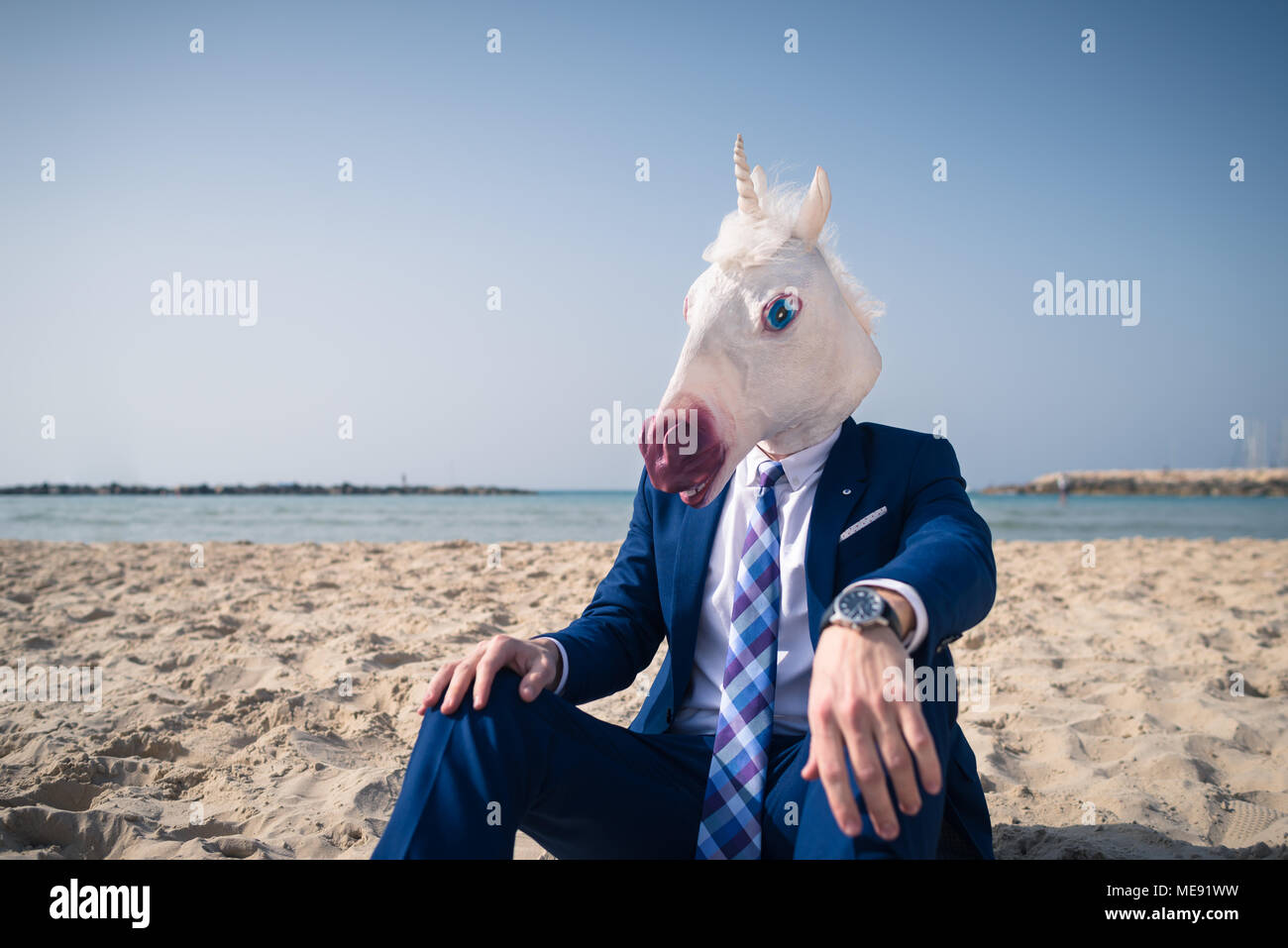 Trendy Mann in lustige Maske sitzt auf dem Sand und genießt Urlaub. Seltsame Mann im eleganten Anzug entspannt am Strand. Einhorn auf Hintergrund von Himmel und Meer Stockfoto
