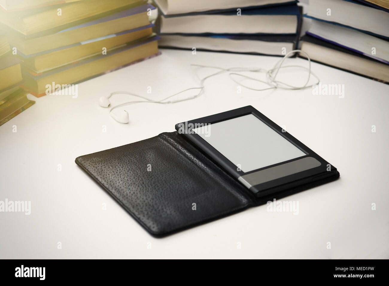 E-Buch liegt auf einem weißen Tisch auf einem Hintergrund von Büchern aus Papier. Kopfhörer liegen in der Nähe. Stockfoto