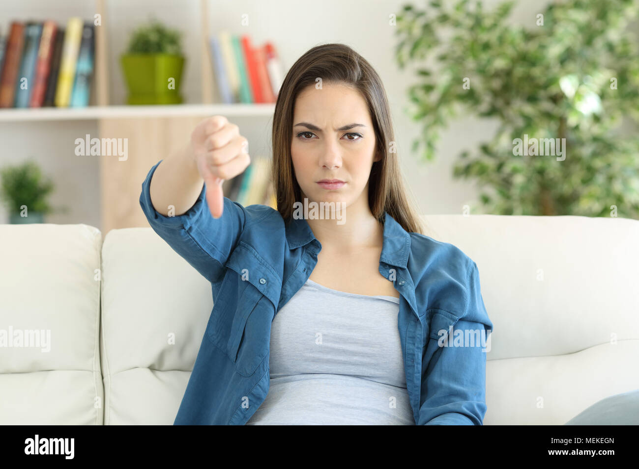 Wütende Frau auf Kamera mit Daumen nach unten sitzen auf einer Couch im Wohnzimmer zu Hause Stockfoto