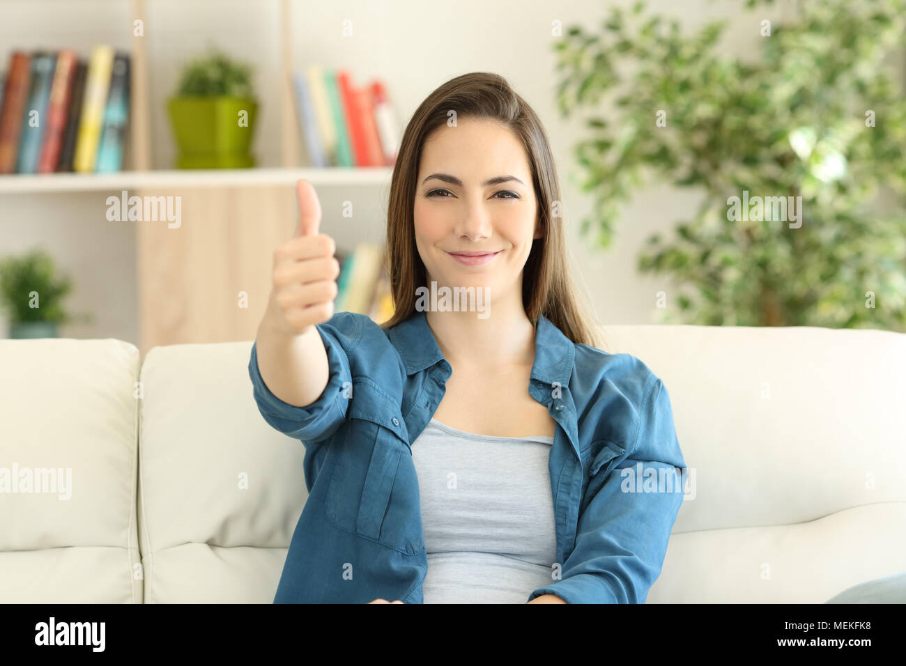 Zufriedene Frau an der Kamera mit Daumen oben auf einer Couch im Wohnzimmer zu Hause sitzen auf der Suche Stockfoto