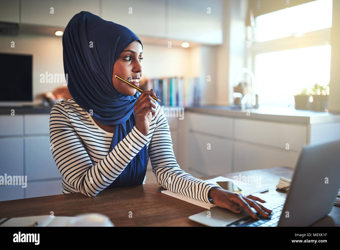 Junge arabische weibliche Unternehmer Tragen eines hijab tief in Gedanken, während an ihrem Küchentisch sitzen, die an einem Notebook arbeitet Stockfoto