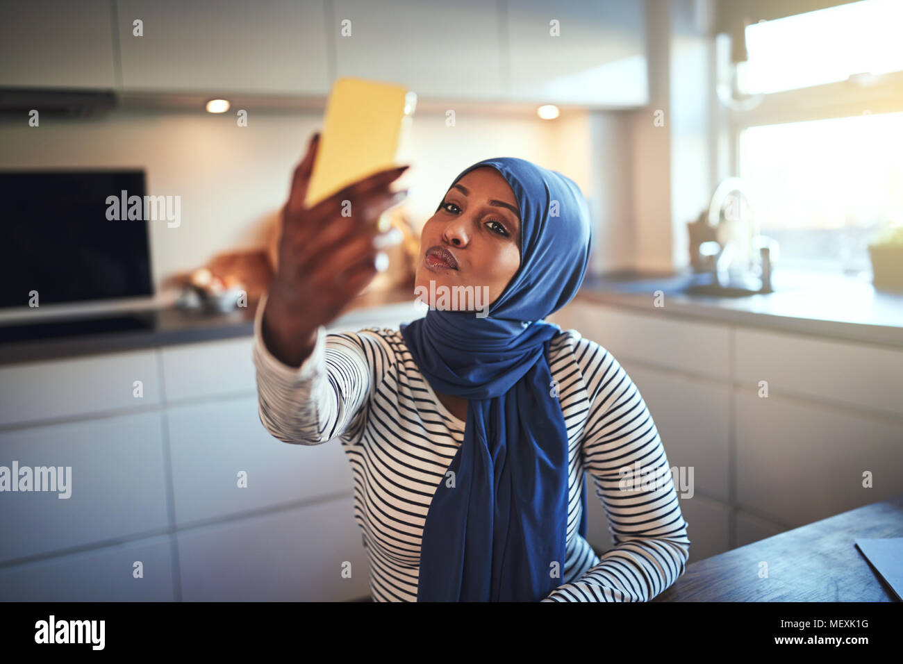 Junge arabische Frau trägt ein Kopftuch sitzen in ihrer Küche zu Hause ein Gesicht während einer selfie mit Ihrem Handy Stockfoto