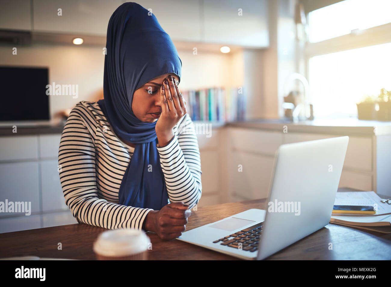 Die junge arabische weibliche Unternehmer Tragen eines hijab an ihrem Küchentisch sitzen, das online arbeitet mit einem Laptop betonte Stockfoto