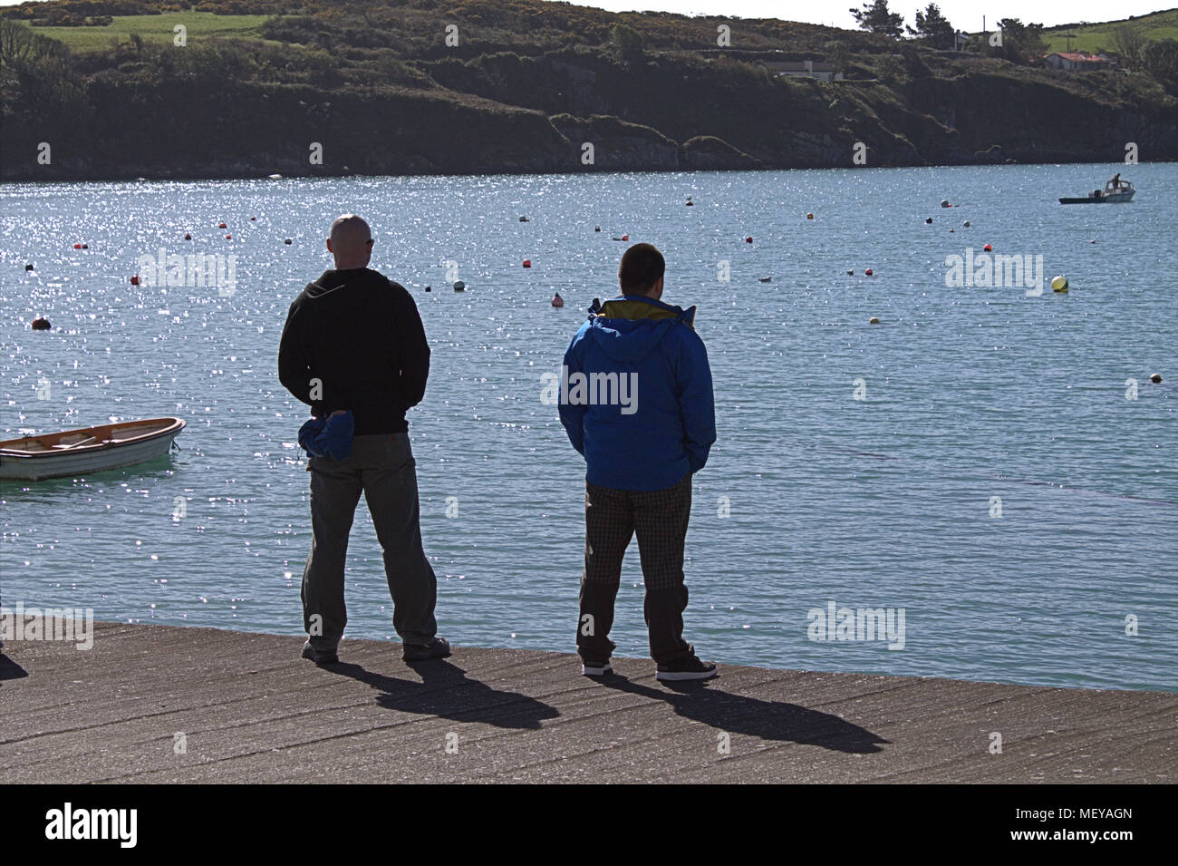 2 im mittleren Alter stehende Männer auf einem Steg im County Cork, Irland beobachten ein Fischerboot in der Ferne. County Cork ein beliebtes Urlaubsziel. Stockfoto