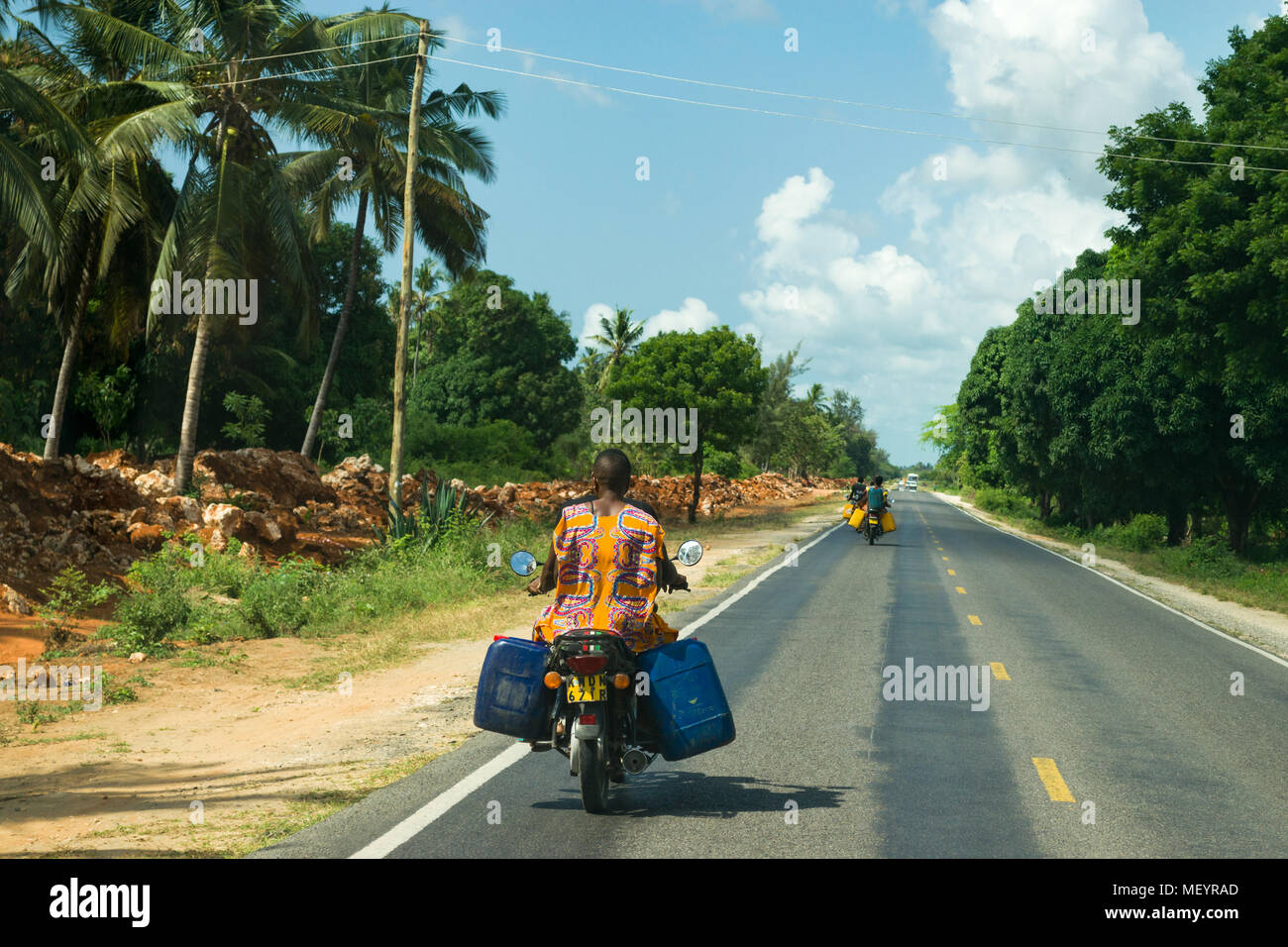 An einem sonnigen Tag in Kenia fährt ein Motorrad mit Fahrgästen, die Wasserfässer tragen, entlang einer Küstenstraße Stockfoto