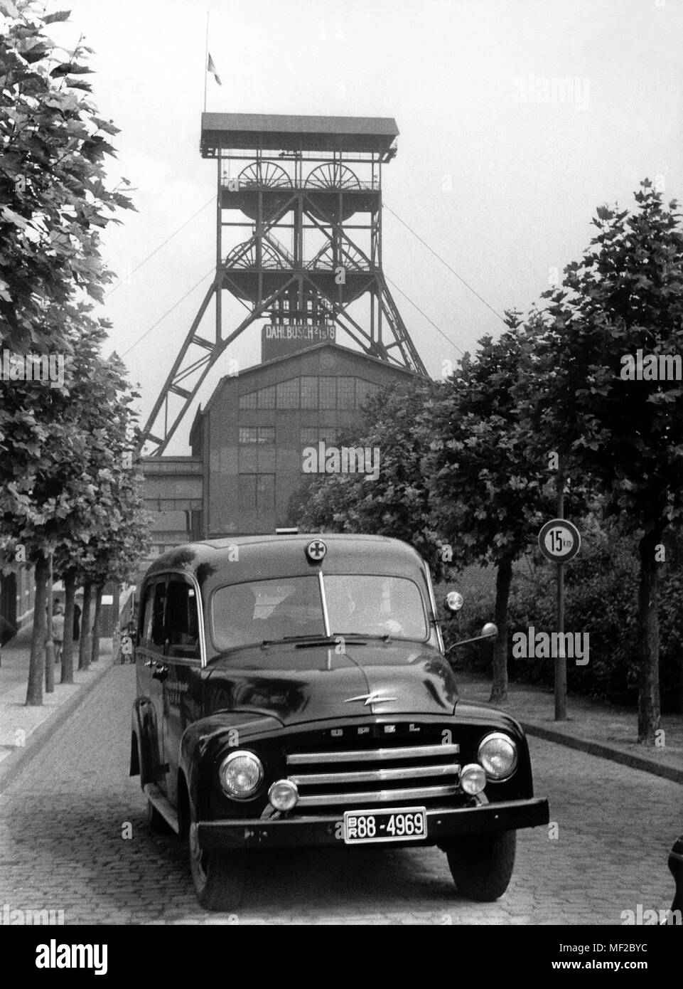 Ein Sanitär Auto verlässt die Zeche. Im Hintergrund die Nachfrage Turm von Schacht Dahlbusch 2/5/8 mit einer Flagge schwenkten auf Halbmast. Am 3. August 1955 ereignete sich auf der Zeche Dahlbusch in Gelsenkirchen zwischen der zehnten und elften Sohle auf der Welle 8 crash Explosion, in denen Insgesamt wurden 41 Bergleute getötet und 21 wurden schwer verletzt. 25 Freunde, die waren in der Nähe der Hochöfen konnte nicht gerettet werden aufgrund der starken mine Brand im betroffenen Strebs. Nach der Rettung arbeiten angehalten wurde, diese Korridore wurden zugemauert, um so eine Brandausbreitung zu verhindern. Bereits im Mai 1955, drei m Stockfoto