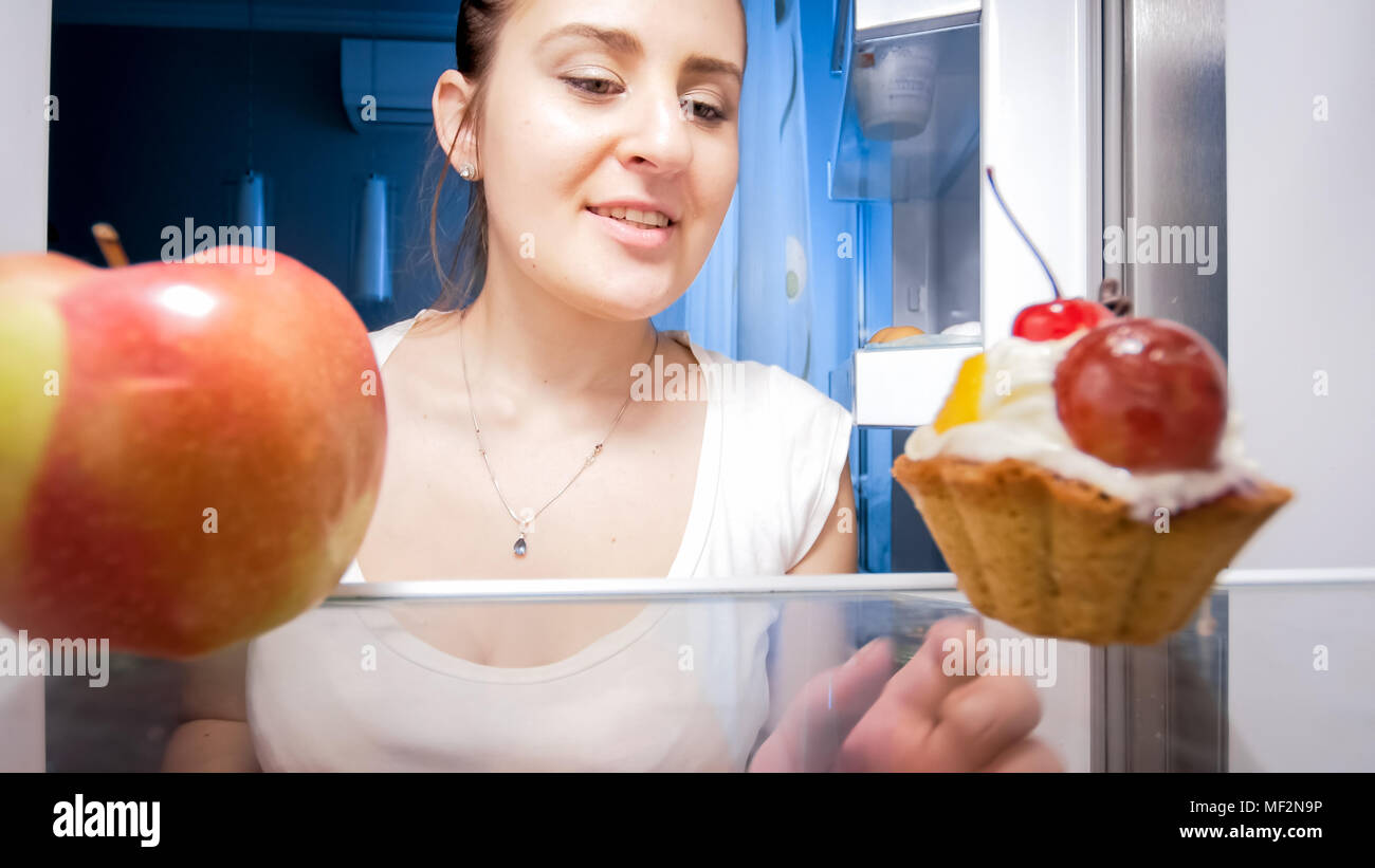 Porträt der jungen Frau zu Zweifeln zwischen Red Apple und süße Kuchen auf dem Regal im Kühlschrank Stockfoto