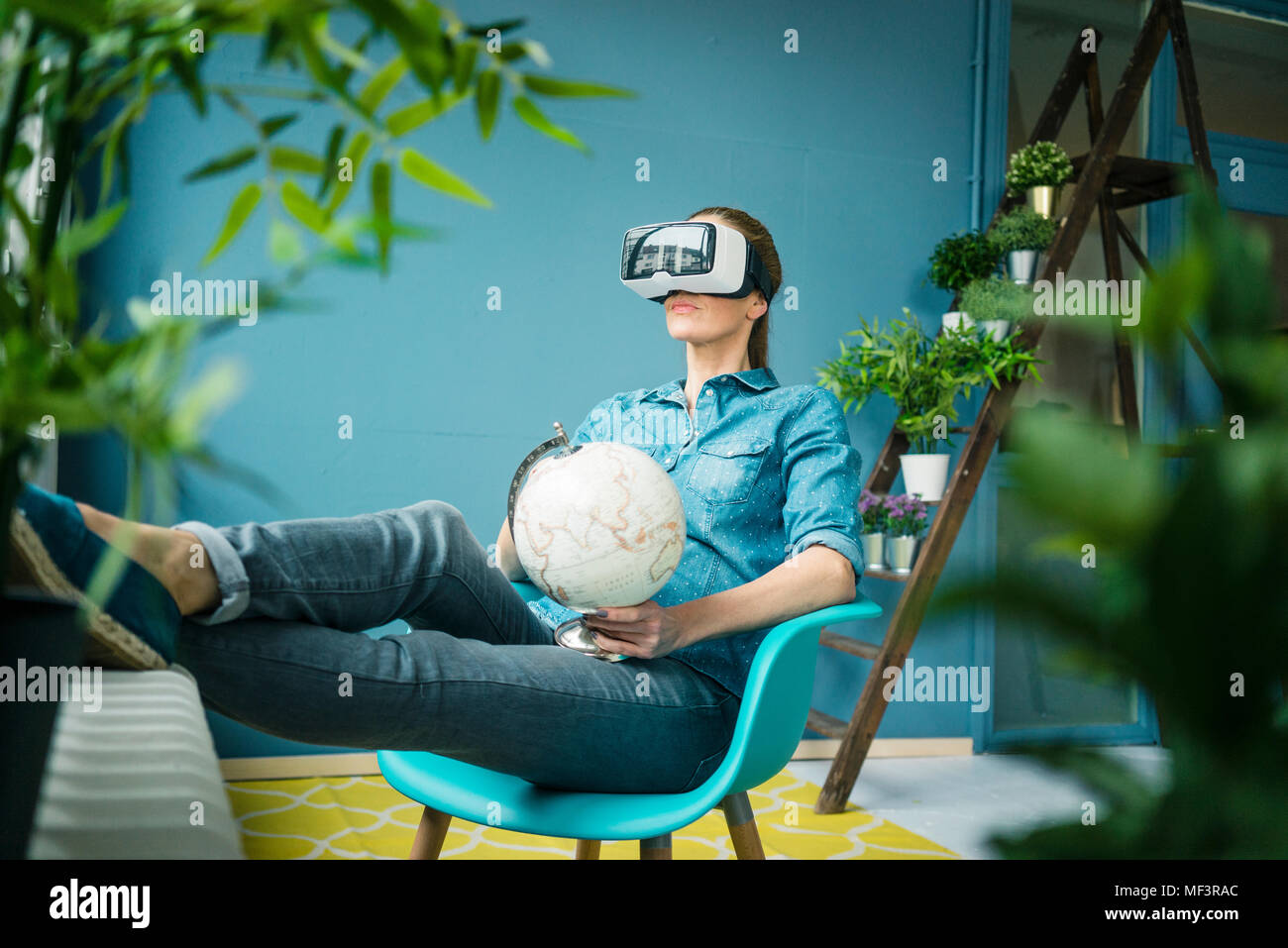 Schöne Frau in ihrem Haus sitzen, mit Pflanzen dekoriert, Globus, VR-Brille tragen Stockfoto