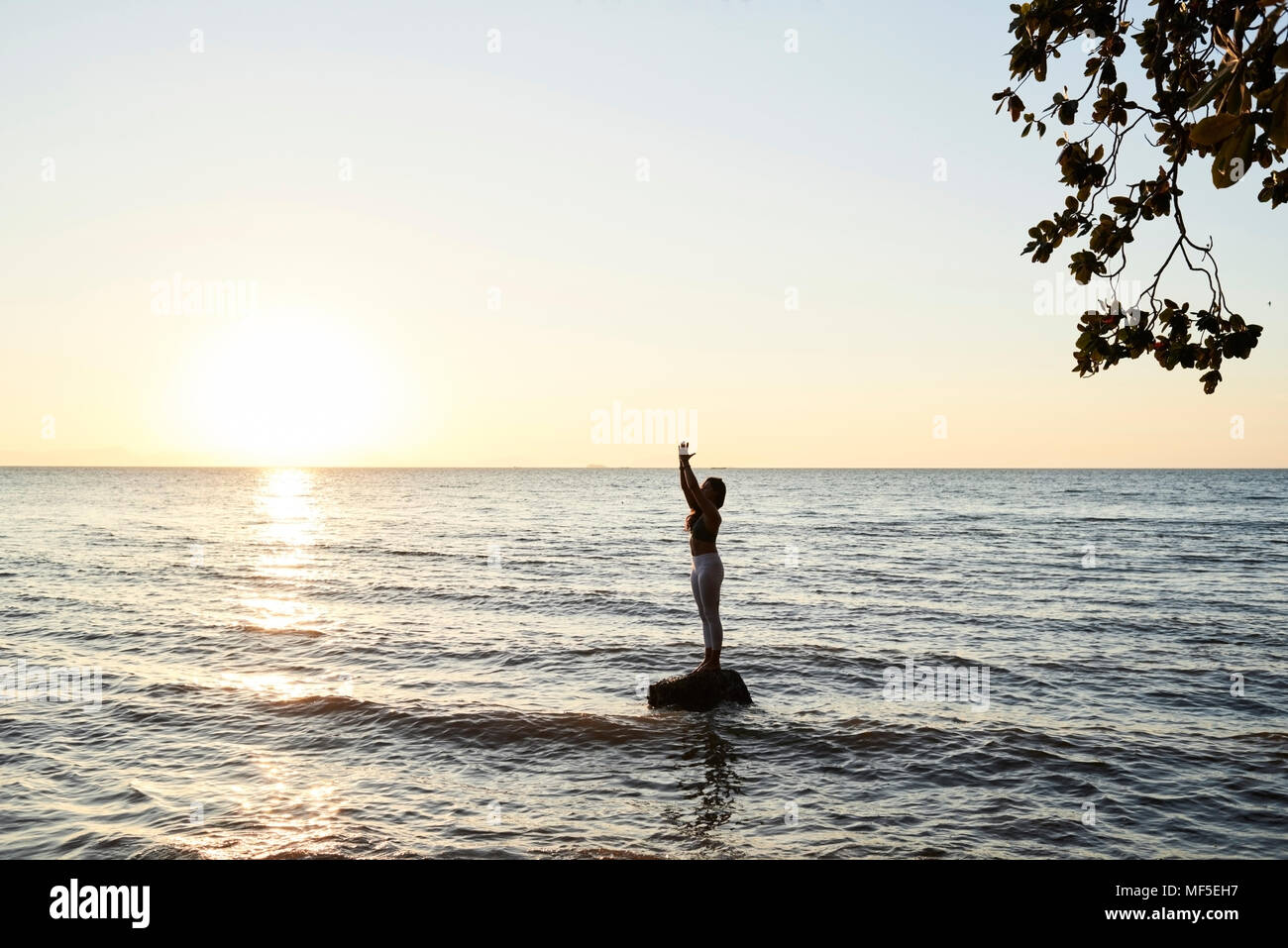 Junge Frau mit Yoga auf einem Felsen im Meer bei Sonnenuntergang Stockfoto