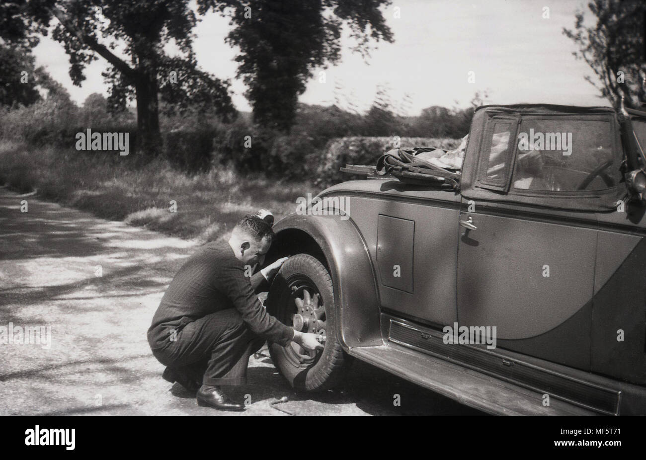 1936, historischen, einen gut gekleideten Herrn im Anzug prüfen einer zurück Reifen auf seinem Zweisitzer oben offenen Auto, auf einem Feldweg in ländlichen Hampshire, England, UK. Stockfoto