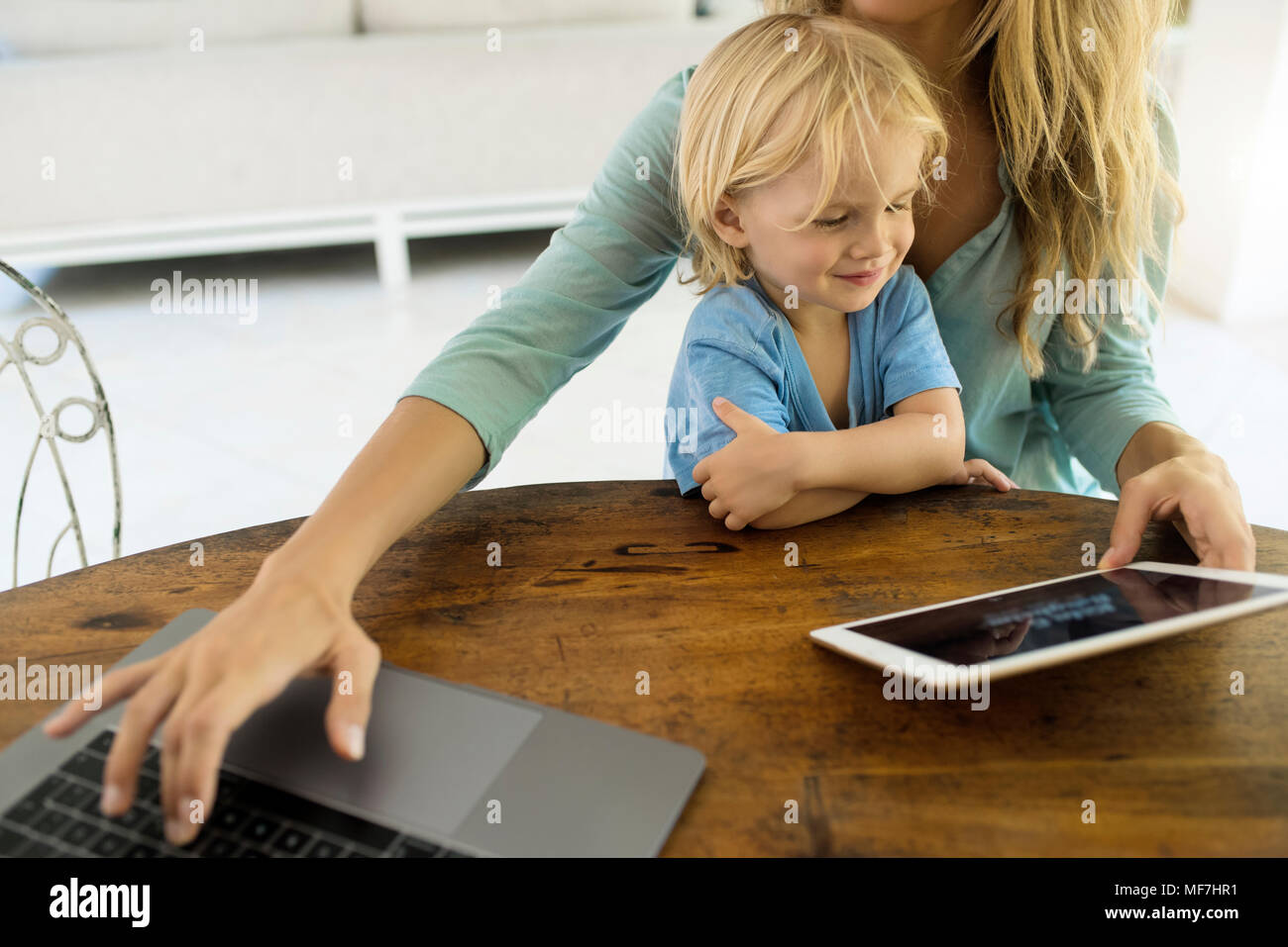 Junge sitzt auf dem Schoß seiner Mutter und mit Blick auf eine Tablette, während seine Mutter arbeitet an einem Laptop ist Stockfoto