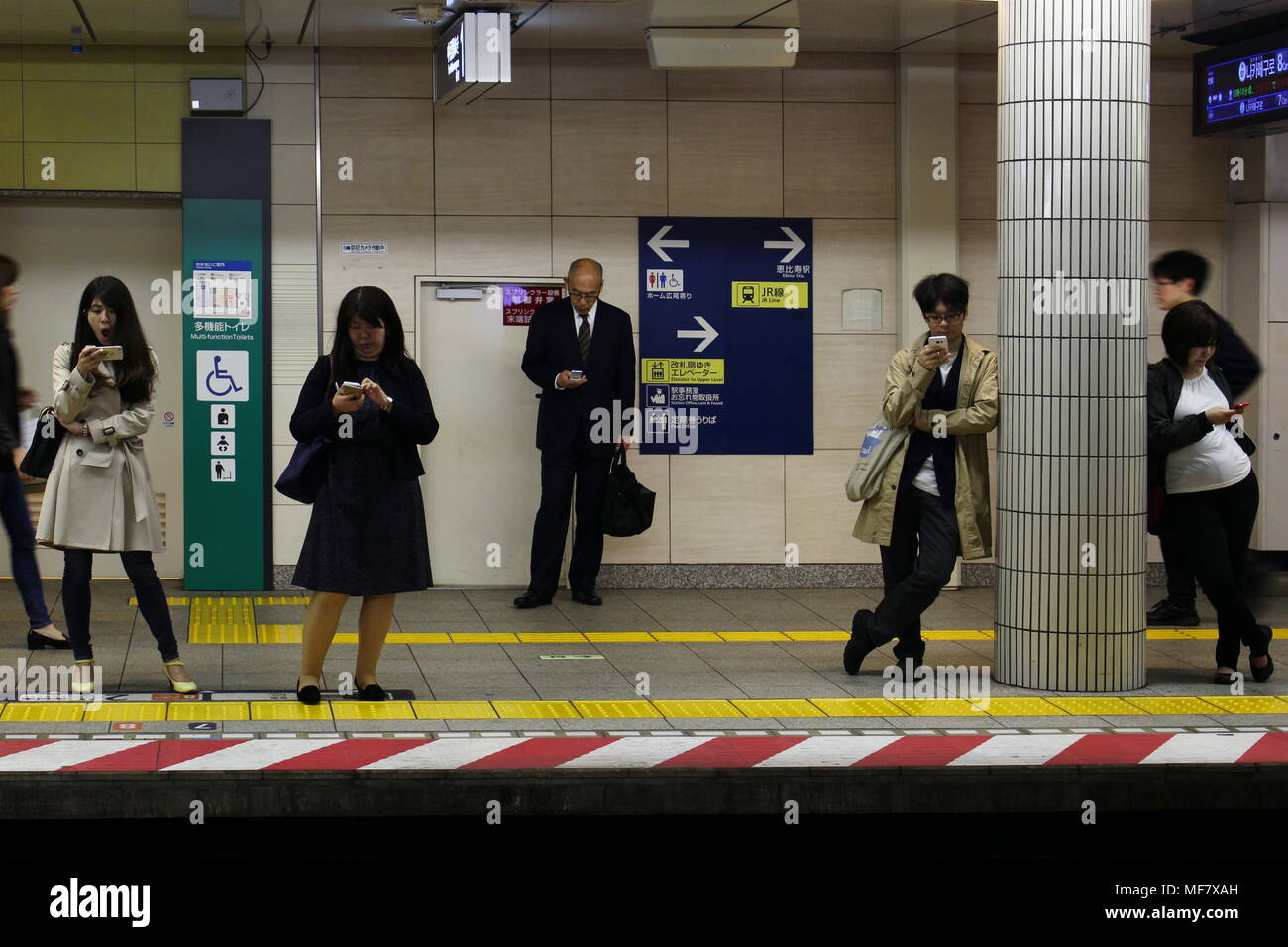 Passagiere auf einer Plattform an der U-Bahn Station Ebisu verwenden ihr Mobiltelefon als Sie darauf warten, dass ein Hibiya Linie Zug zu gelangen. (April 2018) Stockfoto