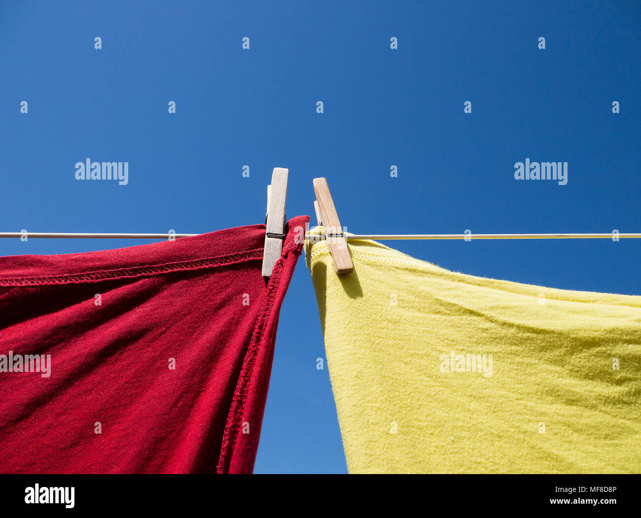 Eine rote und eine gelbe T-Shirt hängen an einer Wäscheleine, gehalten, mit Wäscheklammern aus Holz. In der Farbe von unten geschossen, mit einem klaren blauen Himmel hinter sich. Stockfoto