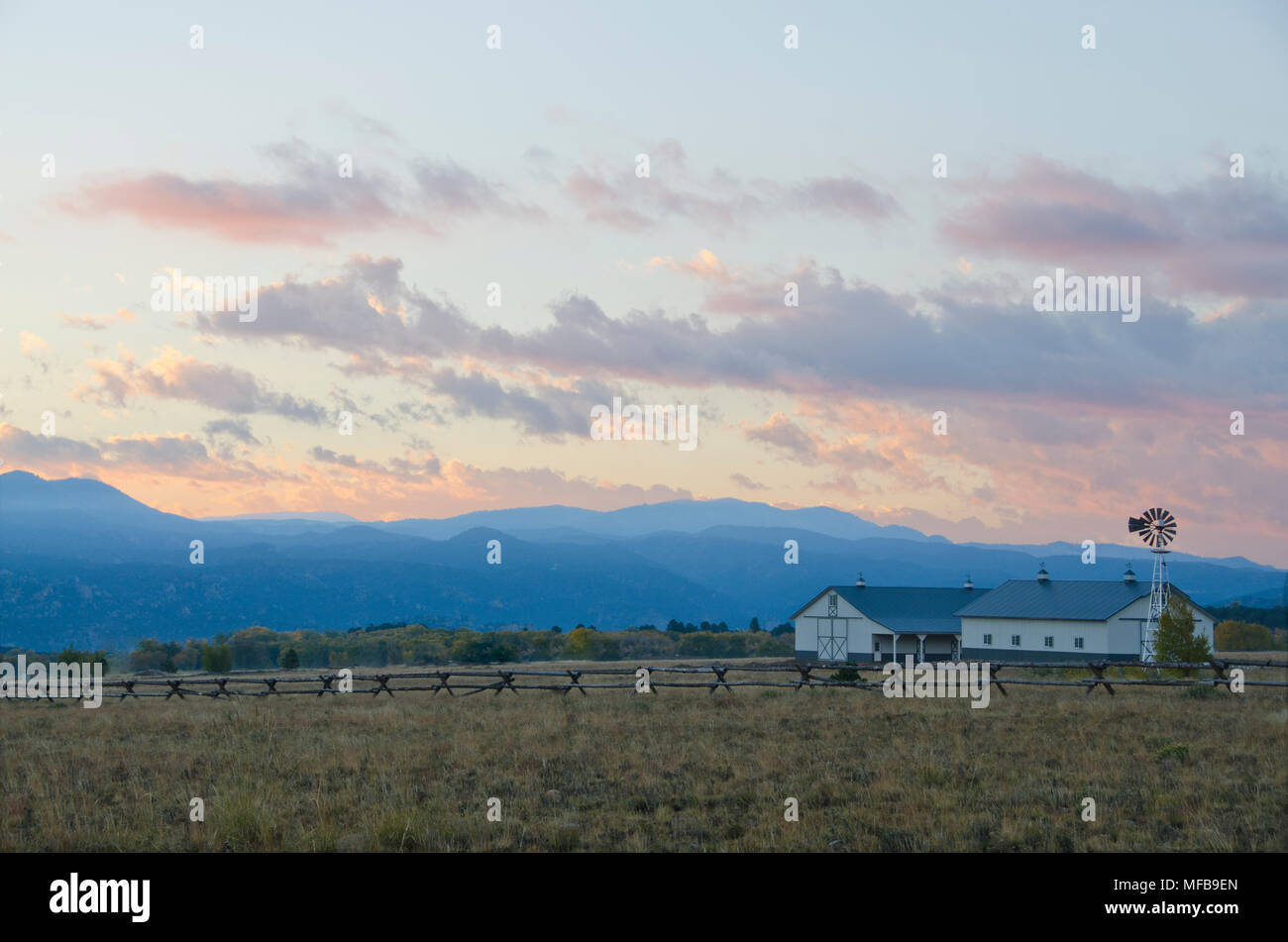 Ein Mountain Ranch, komplett mit Pferdestall und Windmühle hinter einem Zaun, Uhren die Sonne hinter dem Moskitonetz Berge. Stockfoto