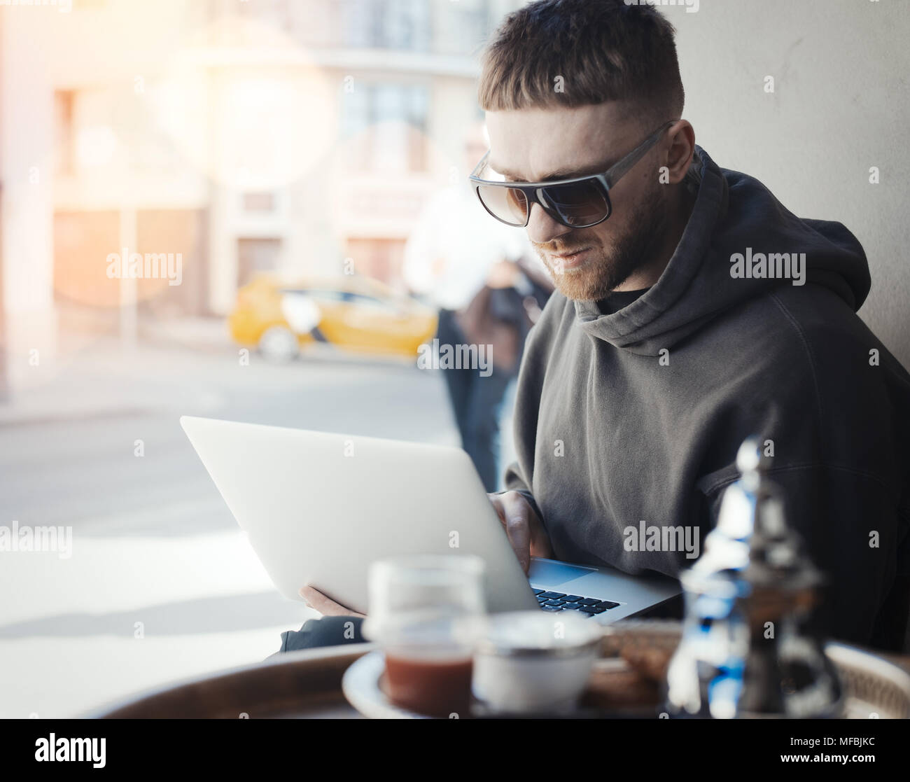 Nachdenklicher junger Mann mit Laptop auf den Knien und an ihm zu arbeiten im Cafe. PC und Mensch stehen im Vordergrund und Hintergrund. Geschirr und Tee sind im Vordergrund Stockfoto
