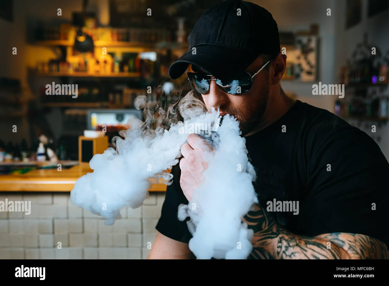 Brutale bärtigen Mann in Sonnenbrille Rauch eine elektronische Zigarette und Releases Wolken von Dampf an der vape Shop. Stockfoto