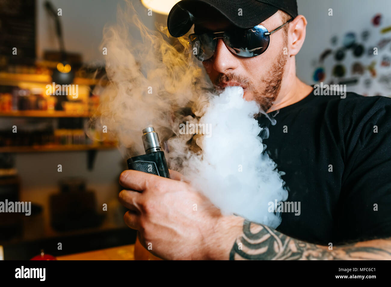 Brutale bärtigen Mann in Sonnenbrille Rauch eine elektronische Zigarette und Releases Wolken von Dampf an der vape Shop. Stockfoto