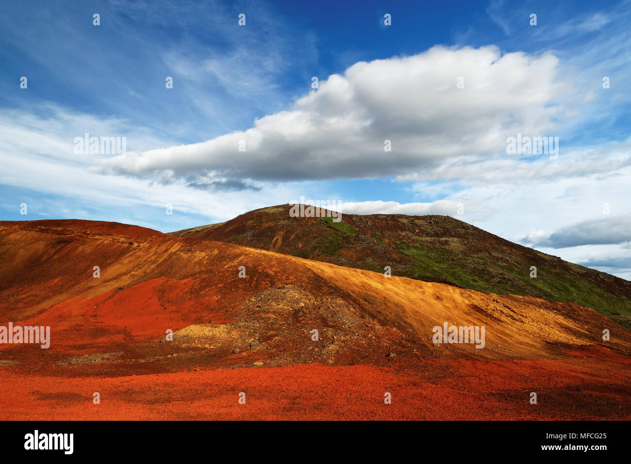 Bunte Ablagerungen vulkanischer Asche in Rot und Gelb auf grünem Hügel, über dem blauen Himmel mit einem markanten Wolkenbildung - Ort: Island, Golden Stockfoto