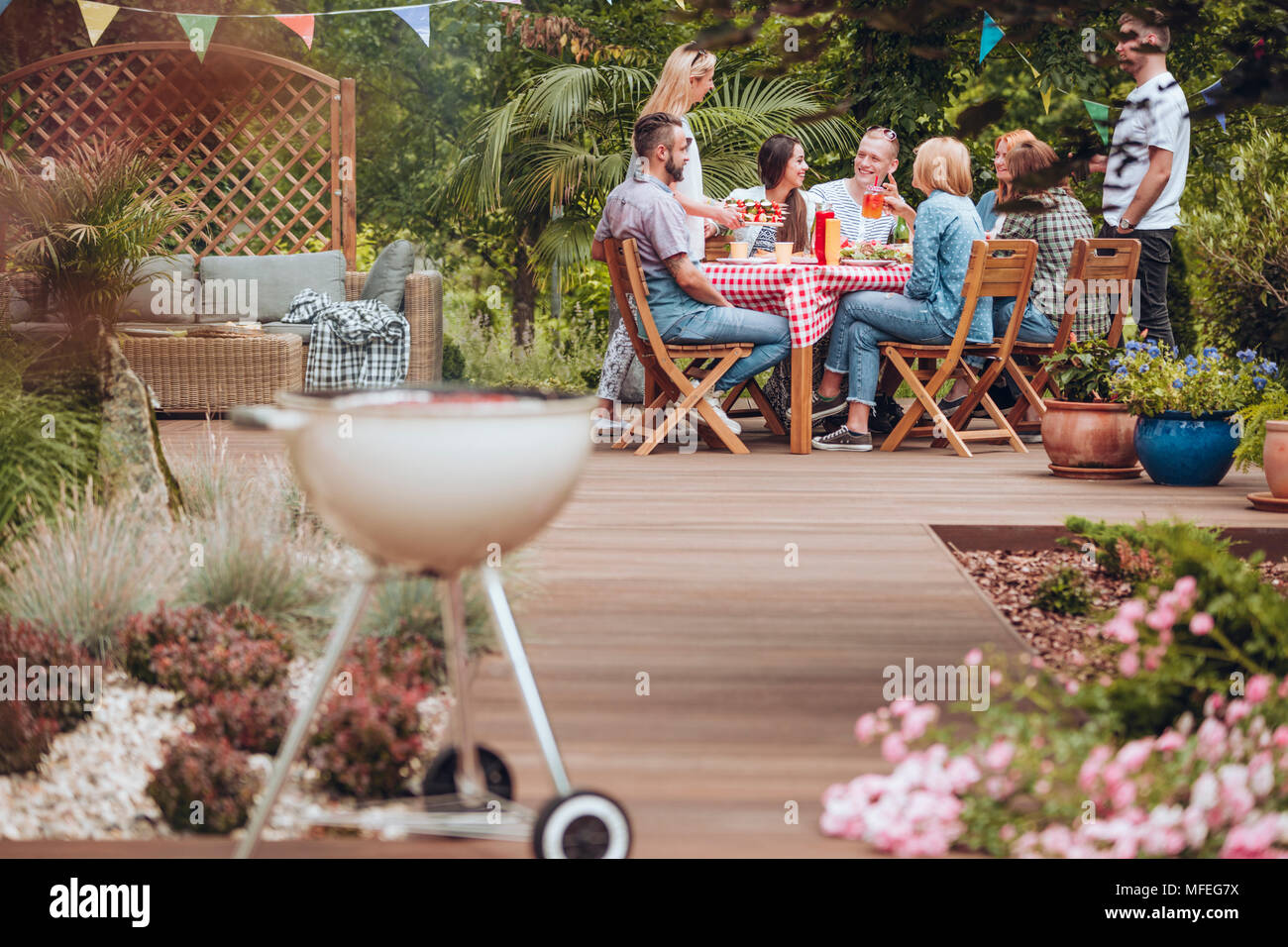 Holz Terrasse im Garten mit einem Grill in der vorderen und glückliche junge Menschen um einen Tisch voller Essen im Sommer bremse Treffen zusammengekommen Stockfoto