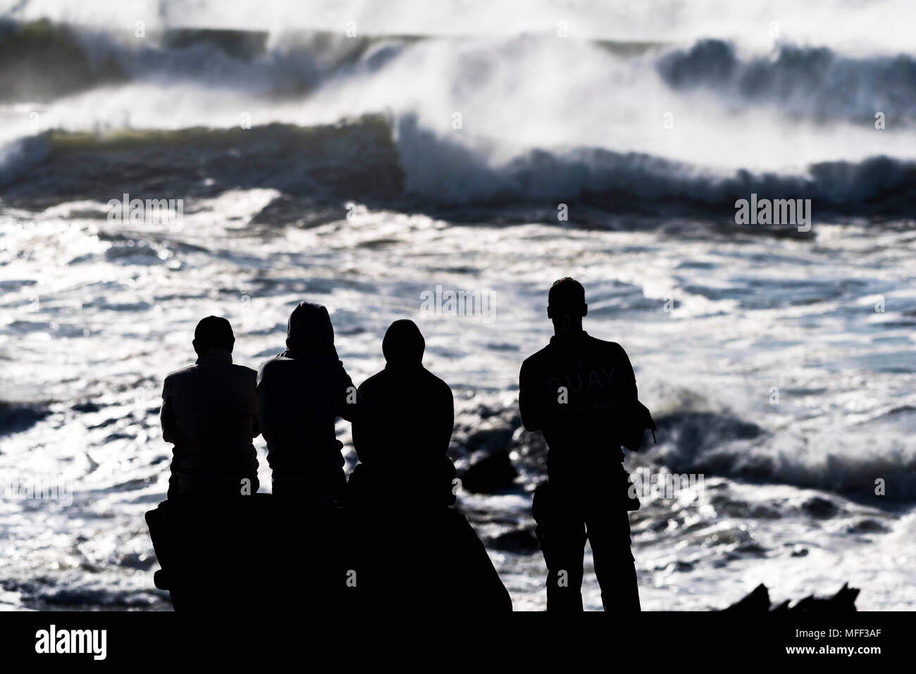 Eine Gruppe von Menschen gegen das helle Sonnenlicht beobachten große eingehende Wellen an der Küste von North Cornwall. Stockfoto
