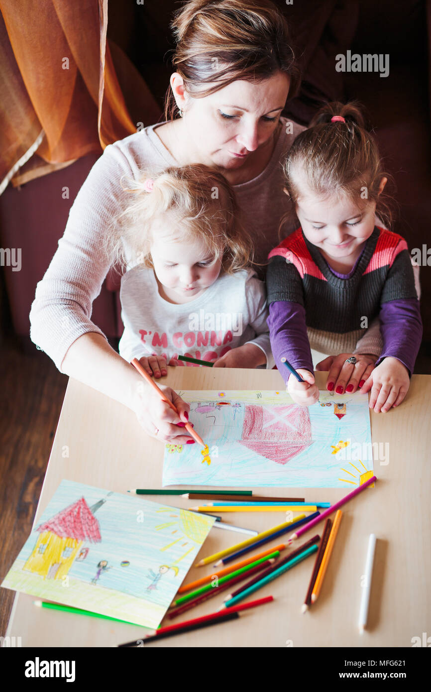 Mama mit kleinen Mädchen Zeichnung eine bunte Bilder von Haus und spielende Kinder mit Bleistift Buntstifte am Tisch drinnen sitzen. Von oben geschossen Stockfoto