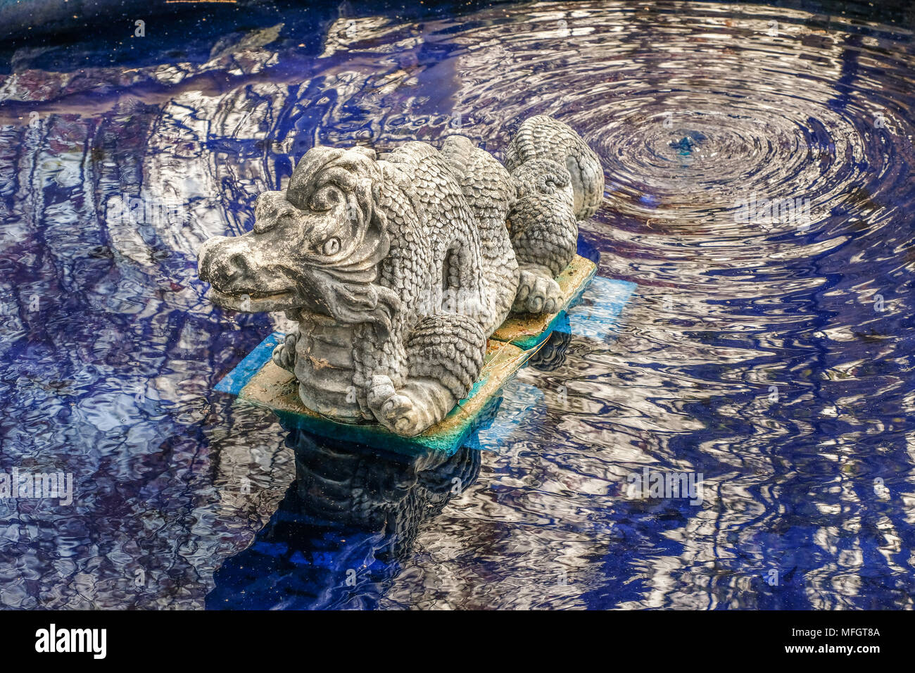 Stein Skulptur eines Drachen in einem öffentlichen Garten; der Drache sitzt in einem Teich mit blau gefärbten Wasser; Reflexionen auf Wasser Stockfoto