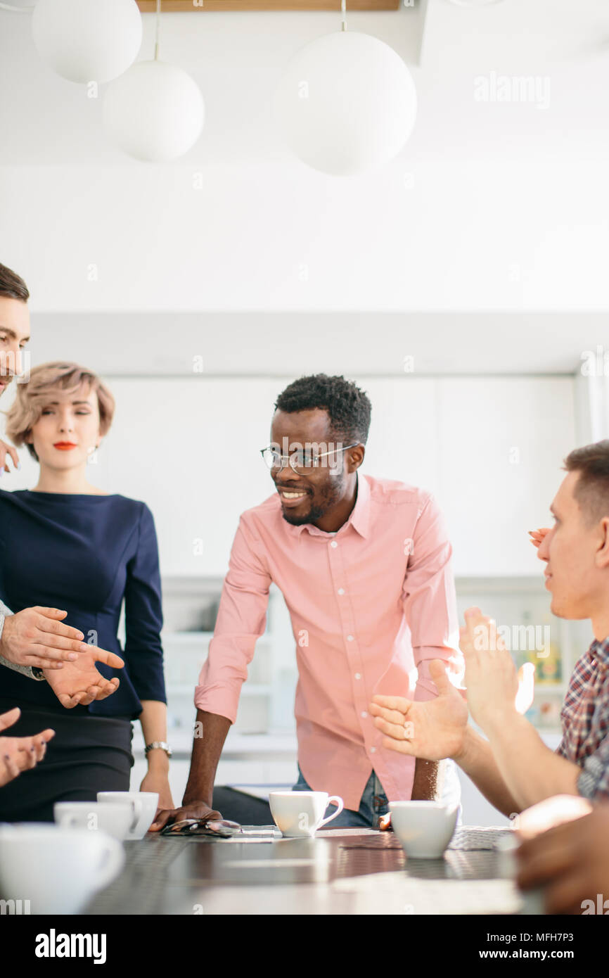 Die enge vertikale Foto von fröhlichen Afrikanischen Manager in rosa Hemd lehnte sich auf den Tisch und sprechen mit den Menschen, während sie Tee trinken. Stockfoto