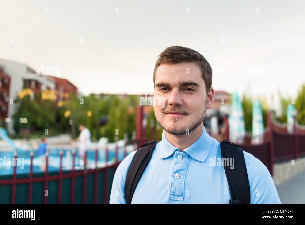 Junger stattlicher Mann mit Schnurrbart steht in der Nähe der Apartmentanlage mit Pool in sonniger Sommertag. Stockfoto