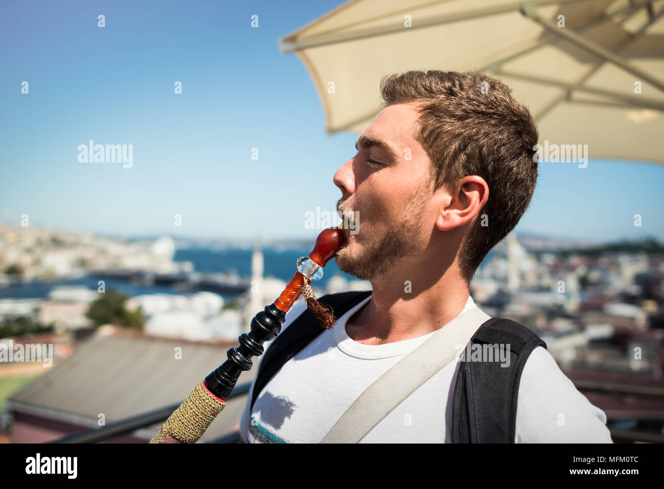 Ein junger stattlicher Mann mit Bart ist das Rauchen einer Wasserpfeife auf der Terrasse mit Blick auf das Stadtbild Stockfoto