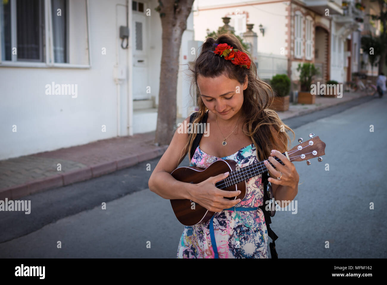 Lächelnde Frau mit Rosen in Frisur hält in den Händen Musikinstrument. Hübsches Mädchen geht auf der Straße und spielt auf der Ukulele. Stockfoto