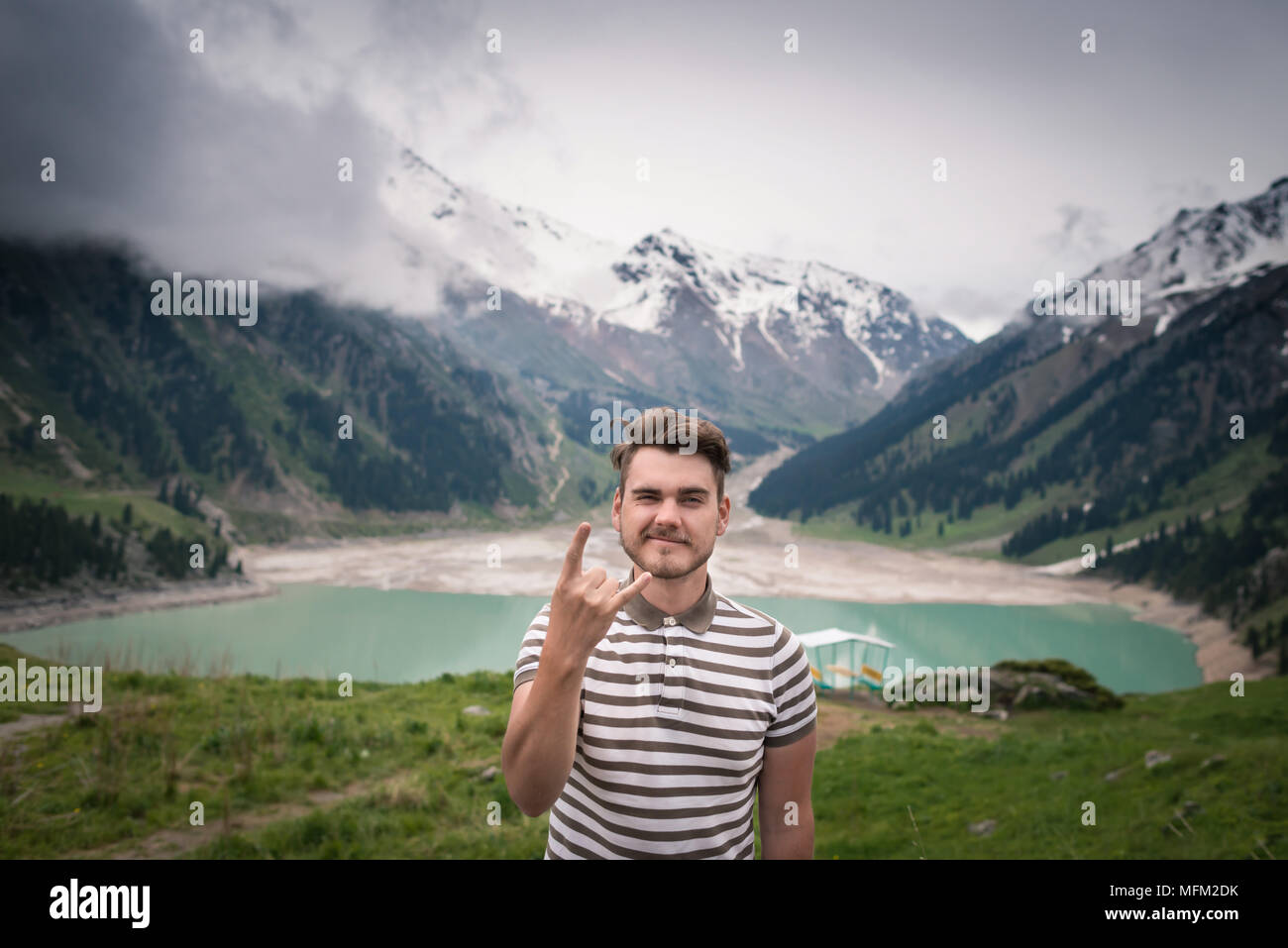Hübscher junger Mann steht auf dem Hügel mit Blick auf den See und einen Rock Geste. Hintergrund mit grünen Berge mit schneebedeckten Gipfeln und Cloud Stockfoto