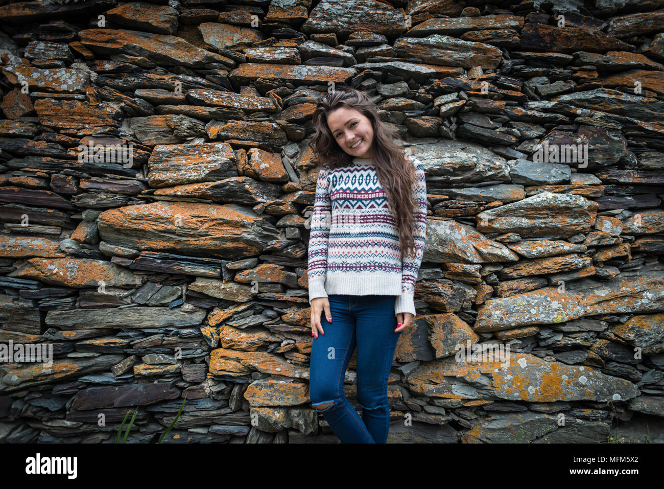 Gerne Frau steht auf isolierten Hintergrund der Steine. Ansicht von vorne. Stilvolle Mädchen in Pullover und Jeans ist weg schauen Stockfoto