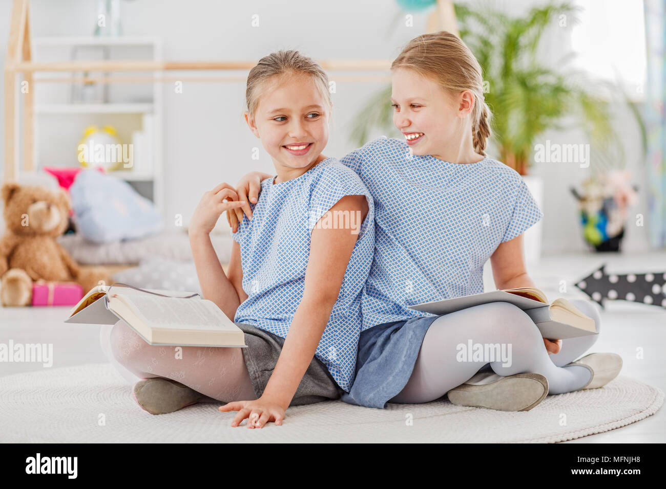 Mädchen lachend mit einem Arm um die Schultern ihrer Schwester, sitzen auf einem weissen Teppich und gemeinsam lesen Stockfoto