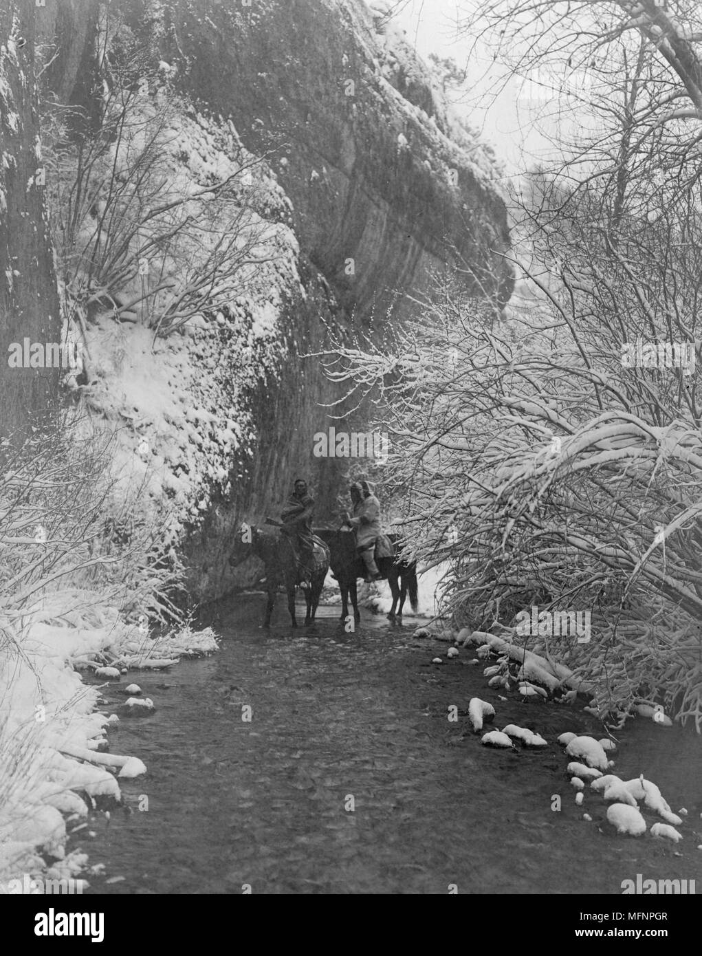 Zwei Crow Indianer auf dem Pferd im flachen Stream von schneebedeckten Bäumen flankiert, unter rock Cliff, c 1908. Foto von Edward Curtis (1868-1952). Stockfoto