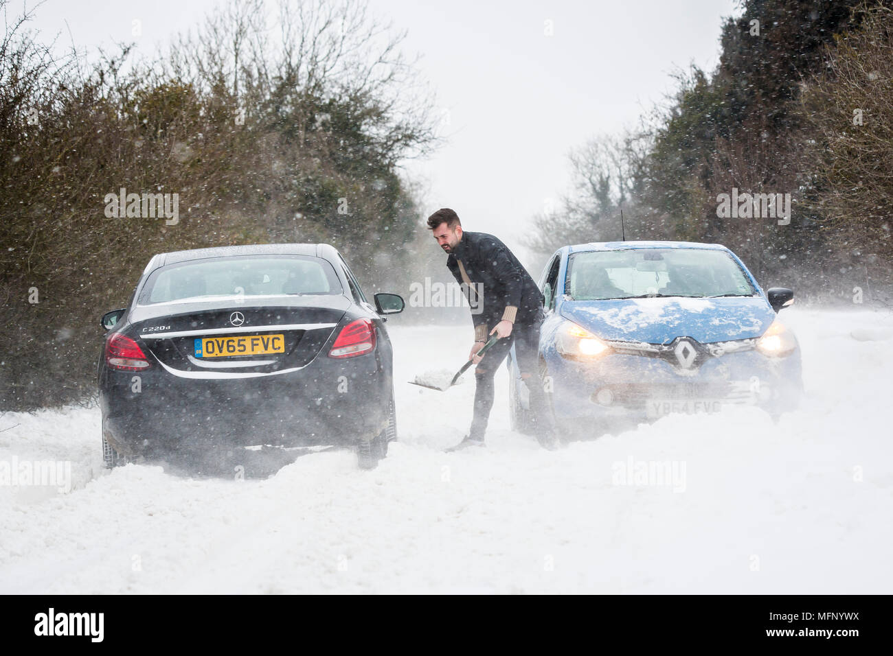 Ein Mann mit einem Spaten sein Auto aus einem Snow Drift steht in tiefem Schnee hat er die starken Winde über dem Land Straße, der wie die anderen Fahrzeuge zu kämpfen, zu graben. Stockfoto