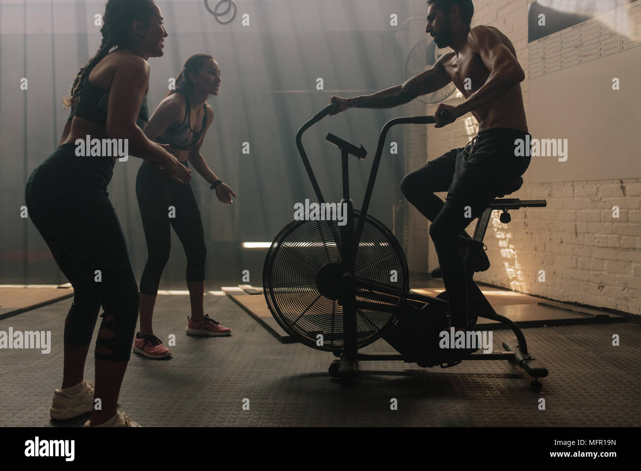 Zwei Frauen motivieren Mann Ausübung auf Air Bike in der Turnhalle. Mann tun, intensives Training im Fitnessstudio Fahrrad mit Frauen. Stockfoto
