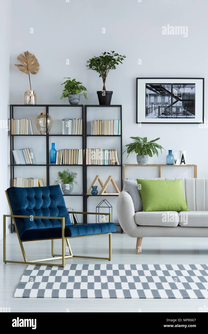 Helles Wohnzimmer mit navy blue, Designer Sessel mit goldenen, Metallrahmen, Glasvasen und Topfpflanzen auf einem Bücherregal Stockfoto