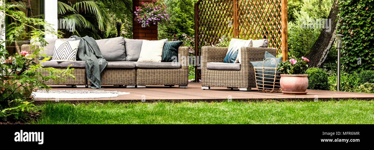 Graue Sofa mit dekorativen Kissen und Körbchen mit Kissen neben Rattan Sessel stehen auf hölzernen Terrasse Stockfoto