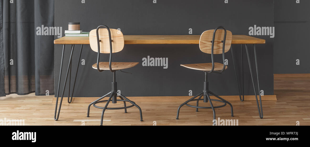 Stühle aus Holz bei langen Schreibtisch gegen schwarze Wand in dunklen Home Office Interior mit Vorhang Stockfoto