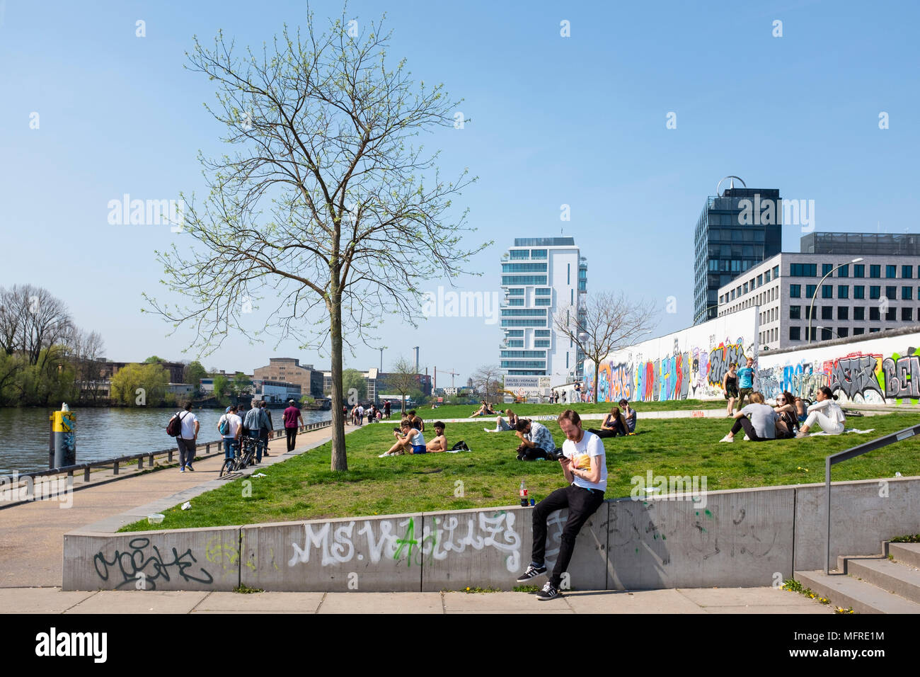 Blick auf Menschen in Riverside Park neben dem ursprünglichen Abschnitt der Berliner Mauer an der East Side Gallery in Friedrichshain, Berlin, Deutschland Stockfoto