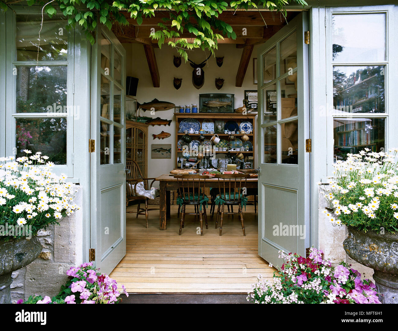 Blumen blühen rund um französische Türen öffnen in ein Land Essbereich mit Esstisch und Stühlen, Holzboden, und eine Sammlung von Geschirr auf offenen shel Stockfoto