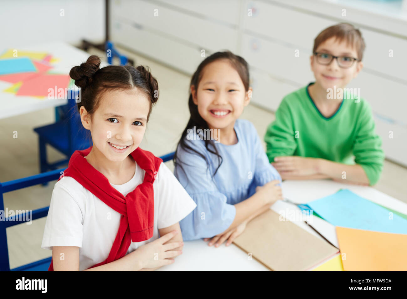 Porträt der Grundschule Kinder mit farbigem Papier am Schreibtisch sitzen und fröhlich lächelnd an Kamera Stockfoto
