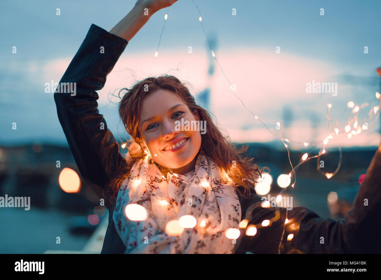 Glückliche junge rothaarige Frau spielen mit Lichterketten im Freien und Lächeln, Blaugrün und Orange Style Stockfoto