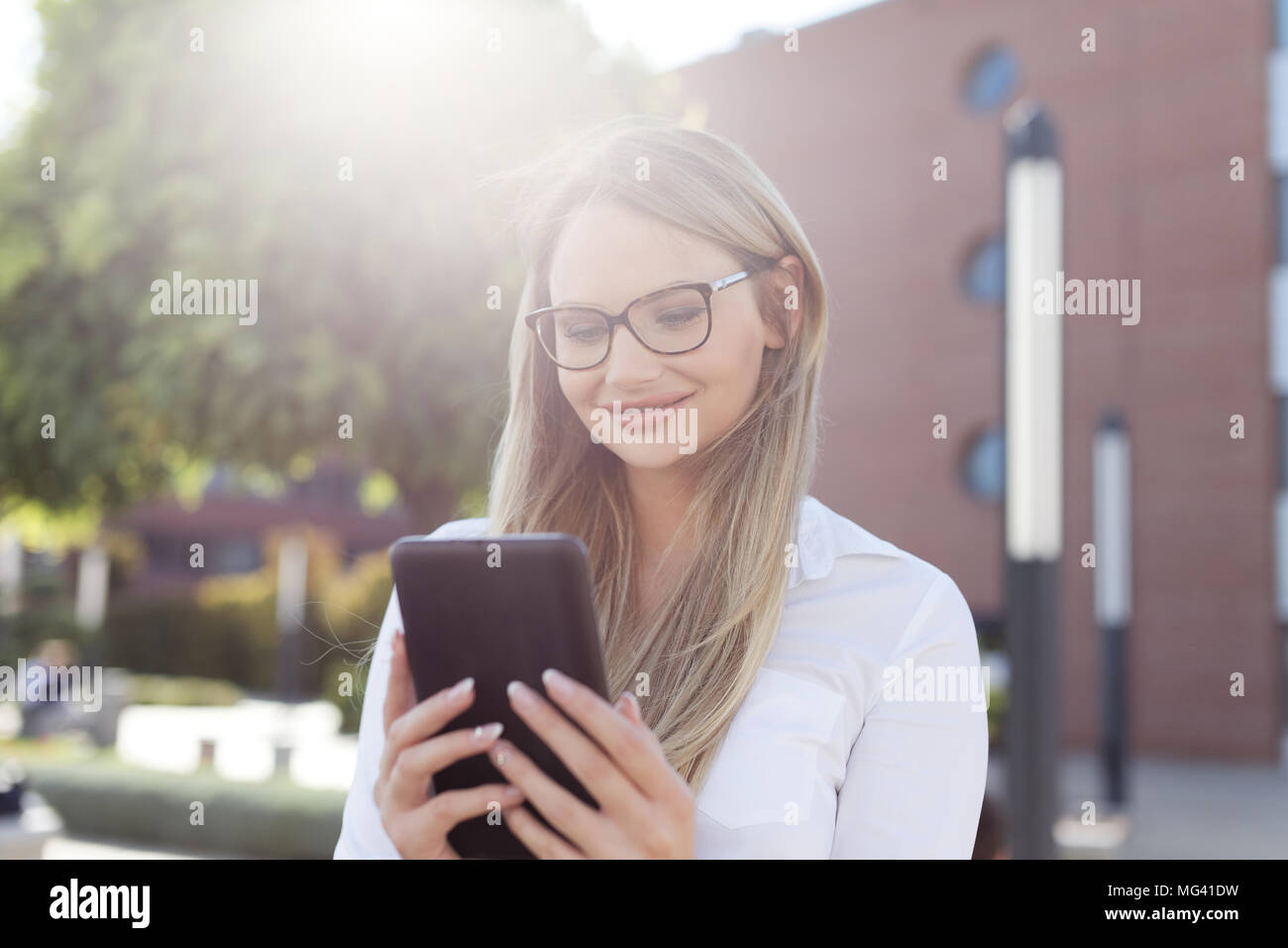 Junge intelligente Blondine Geschäftsfrau in Brillen lesen Nachricht auf Tablet im Freien, mit Sonnenlicht Stockfoto