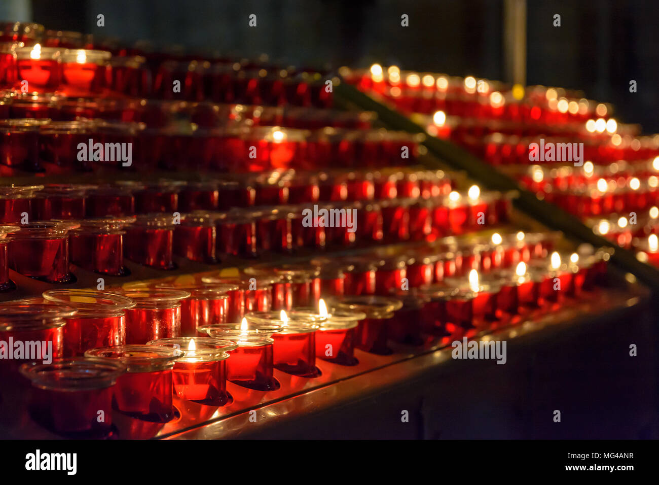 Kleine weiße Votiv Kerzen beleuchtet und in rotem Glas Leuchter unter Leeren platziert, aufgereiht auf einem Kerzenhalter stand in einer katholischen Kirche. Stockfoto