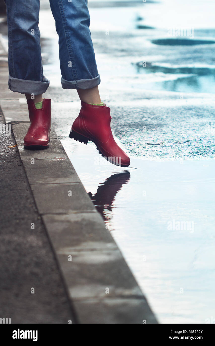 Seitenansicht Nahaufnahme des jungen kaukasischen Frau Füße mit Rot kurze Stiefel und Jeans auf Bürgersteig und tritt in eine Pfütze von Wasser af Stockfoto