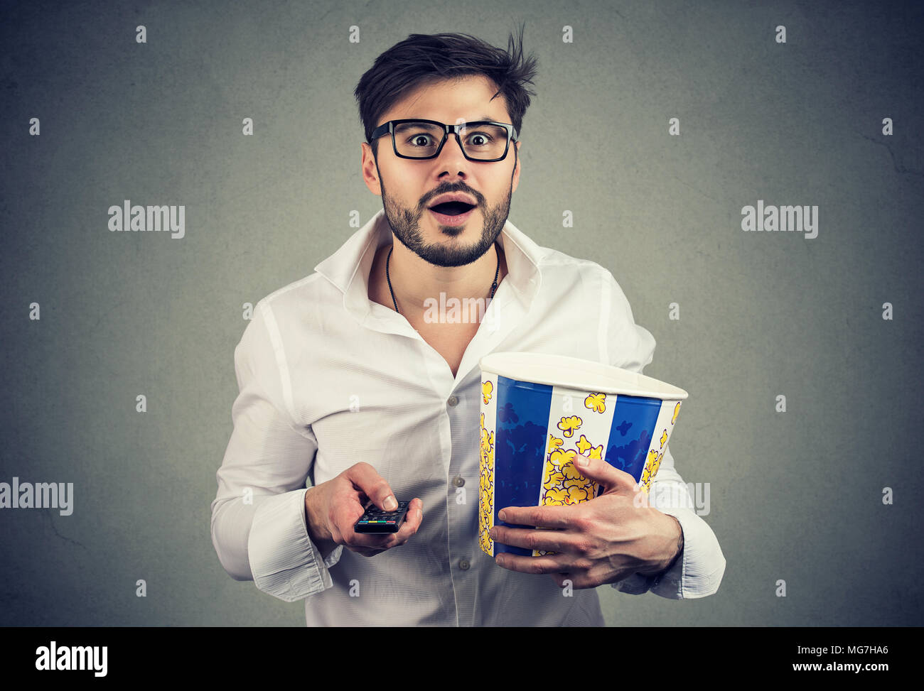 Gut aussehender bärtiger Mann in Gläsern holding Eimer Popcorn und Fernbedienung Fernsehen mit Erstaunen. Stockfoto