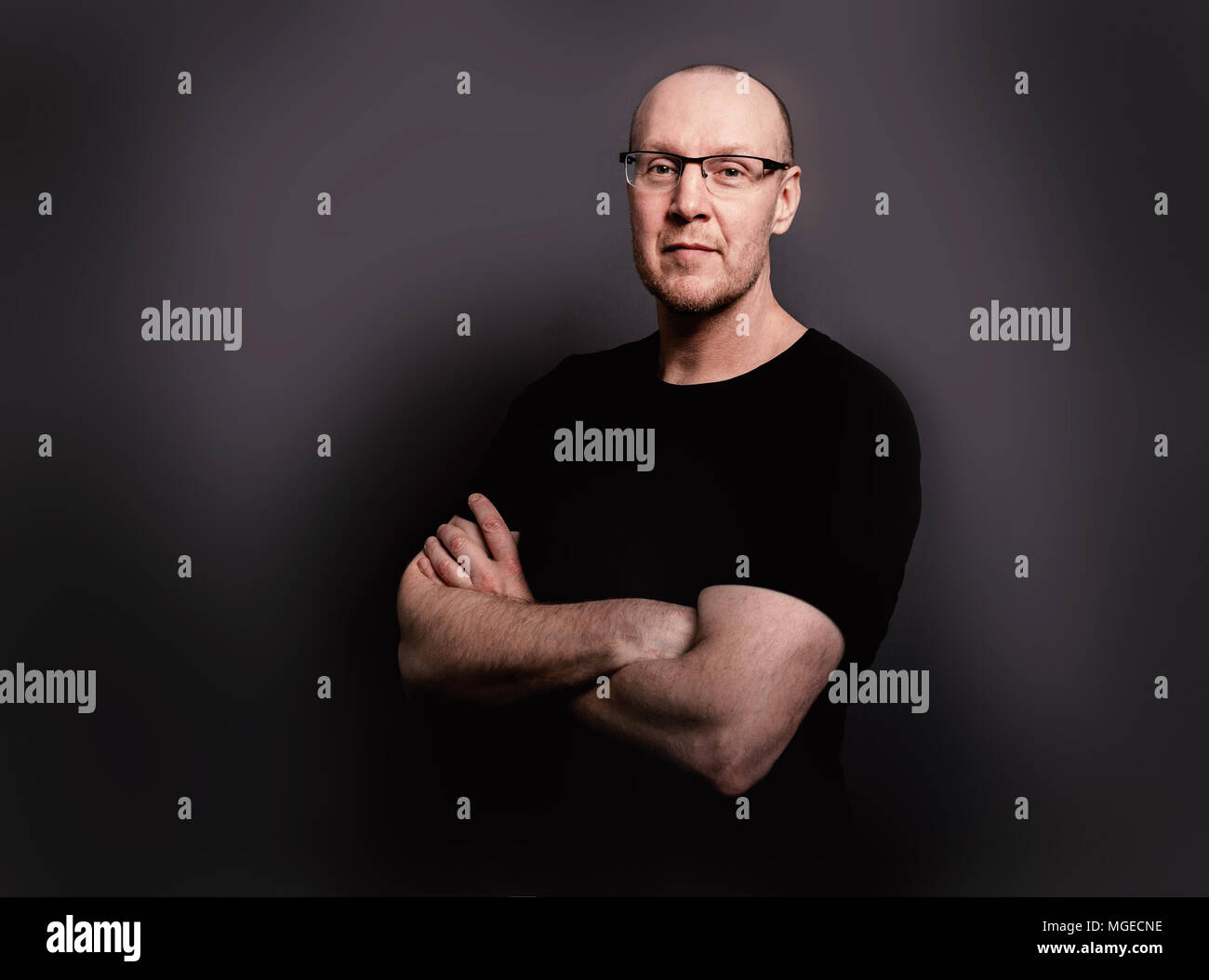Studio Portrait einer mittleren alter Mann mit Brille und lächelnd mit Waffen in ein schwarzes T-Shirt kreuzte vor einem schwarzen Hintergrund in Großbritannien Stockfoto