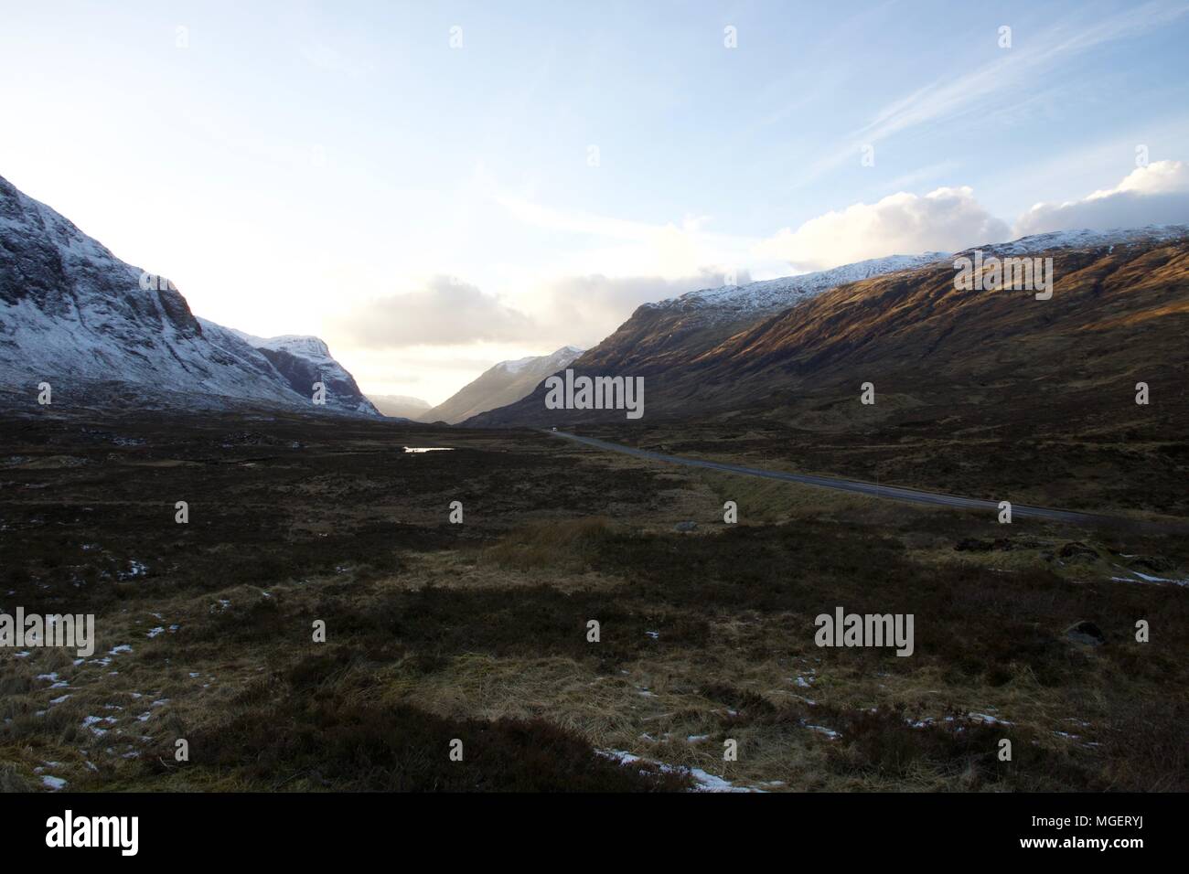Eine konkrete Straße kreuzt die schottischen Highlands bei Sonnenuntergang mit schneebedeckten Gipfeln und typischen Herbstfarben am unteren Rand des Tals Stockfoto
