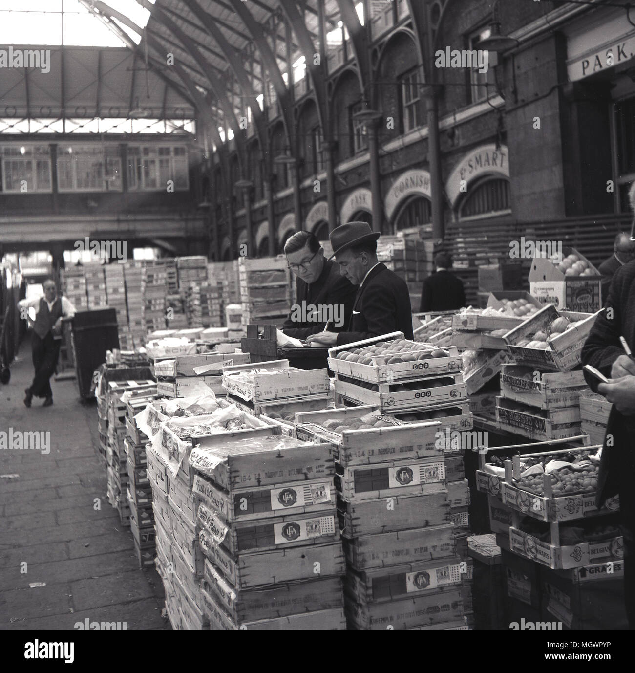 1950er Jahre, historische Bild, zwei Händler machen Sie sich Notizen über ihre Verkäufe, die durch ihre Kisten mit Obst in der Halle am berühmten Obst- und Gemüsemarkt in Covent Garden, London, England, UK umgeben. Stockfoto